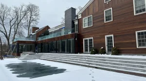 A modern two-story house with dark wooden siding and large windows, situated in a snowy landscape.